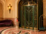 Ornate green and brass elevator in an elegant hotel lobby with polished pink marble floors and a purple settee.