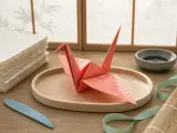A coral paper origami crane resting on a wooden tray beside a stack of textured paper sheets and crafting tools.