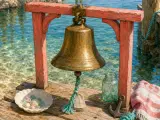 A brass bell hung from a weathered red wooden frame on a pier overlooking clear turquoise sea water.