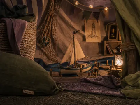 A toy sailboat docked at a miniature wooden pier lit by a small lantern inside a cozy blanket fort.