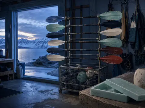 Kayak paddles on a metal rack inside a boathouse looking out at snow-covered mountains across a fjord.